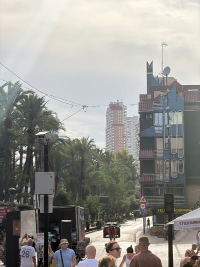 The image depicts a street scene in Benidorm, Spain, likely along a busy thoroughfare lined with palm trees and multi-story buildings. In the foreground, several people are visible. To the left, a man wearing a white t-shirt with "ENGLAND 26" is being photographed. Adjacent to him, a man in a blue t-shirt and a hat is looking towards the right. Further to the right, a group of people are gathered, some appearing to be seated outdoors under a white awning.

In the middle ground, a red car is driving down the street, and a person is riding a bicycle in front of it. Traffic signals and street signs, including a "10" speed limit sign and a bicycle lane sign, are present. A delivery truck, partially open and loaded with cases of bottled water, is parked on the right side of the road.

The background features tall, modern buildings, characteristic of Benidorm's skyline, including a distinctive blue and red multi-story structure with balconies and curved window designs. A security camera is mounted on a pole in the left-center of the image, amidst the palm trees. The sky is overcast, suggesting a hazy or cloudy day. The overall impression is a lively urban environment with both residential and commercial activity.