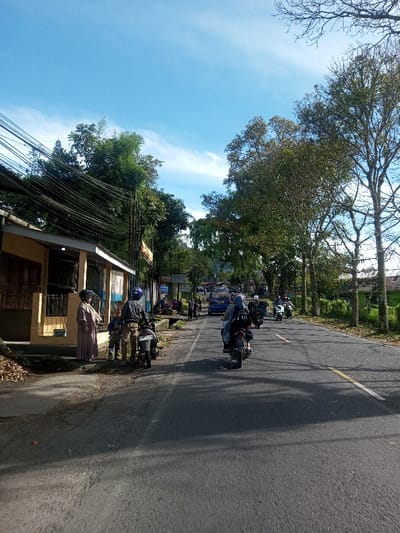 The image captures a daytime street scene in Phonengam, Laos. A two-lane asphalt road, marked with a double solid yellow line and a solid white edge line, recedes into the distance under a bright blue sky with wispy clouds.

On the left side of the road, a single-story building with a light yellow facade and blue trim is visible, partially obscured by a dense tangle of overhead electrical and communication wires. In front of this building, on a dirt and concrete shoulder, two individuals in light brown long dresses, possibly with head coverings, stand facing each other. To their right, a third person wearing a blue helmet and dark patterned shirt stands next to a parked white motorcycle, facing away from the camera. Another individual in a dark shirt and light pants is partially visible near this motorcycle.

On the road, multiple motorcycles are traveling away from the camera. The closest motorcycle carries two riders, both wearing helmets; the rear passenger is wearing a grey hijab and a black backpack. Further down the road, additional motorcycles are visible, along with a blue van.

The right side of the road is lined with a row of mature trees featuring green foliage, some with visible bare branches. Sunlight casts distinct shadows across the road from these trees and the moving vehicles. In the background, more dense greenery suggests a forested area or hills.