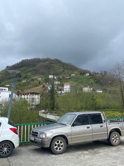 The image shows a hilly landscape with scattered buildings, characteristic of a mountainous region. A silver pickup truck is parked on the roadside, partially obscuring the view of the houses on the hillside. In the foreground, there is a white car parked beside the truck, and a sign with the inscription "KIKI" can be seen. The sky is overcast, suggesting a cloudy day. Given the reference to Kemalpaşa, Turkey, it's reasonable to assume that this scene is located in or near that city.