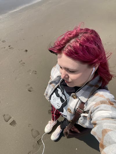 An individual with bright magenta hair is depicted from an overhead perspective, walking or standing on a sandy beach in Ulcinj, Montenegro. The person wears a plaid button-up jacket in shades of light grey, white, and tan over a black t-shirt with a white graphic of a female cartoon character wearing headphones. White wired in-ear headphones are in place. Light pink canvas sneakers are visible on the feet, and a dark brown leather shoulder bag is worn across the body. The individual is looking downwards. The beach consists of dark grey, fine-grained sand, exhibiting numerous footprints. The edge of a body of water, consistent with a sea or ocean, is visible in the upper left, showing small waves along the shoreline. The scene is illuminated by clear daylight.