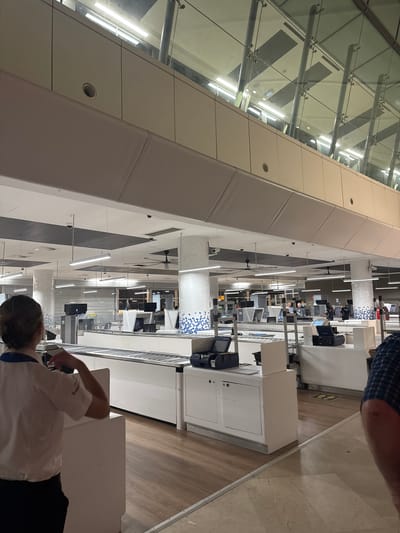 The image depicts a domestic airport terminal in Manises, Spain, specifically the check-in area. In the foreground, a female airport employee, wearing a white collared shirt and black trousers, stands with her back to the camera, operating a check-in counter. The counter features a conveyor belt, a computer monitor, and a scanner. Behind her, several other check-in counters are visible, some occupied by staff and passengers. The terminal features modern architecture with white tiled pillars, fluorescent lighting, and large glass windows. The environment appears clean and orderly, with signage and safety markings indicating social distancing protocols. The overall atmosphere is that of a busy but functional airport.