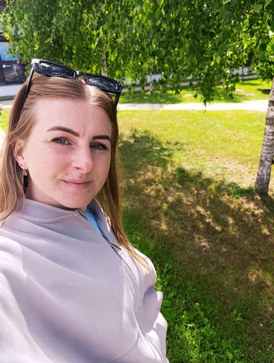 The image is a selfie taken outdoors on a sunny day. A young woman with shoulder-length blonde hair, freckles, and blue eyes is the main subject. She is wearing a light gray zippered jacket over a light blue shirt and black-framed sunglasses perched on top of her head. She has a small hoop earring with a square charm in her left ear. Her expression is a gentle smile, and she appears relaxed and happy.

The background consists of a lush green lawn with dappled sunlight and shadows cast by the leaves of a birch tree. A pathway is visible in the distance, bordered by more green grass. The overall setting suggests a park or a similar outdoor recreational area. The time of day is likely midday or early afternoon, given the bright sunlight and the angle of the shadows. The weather is warm and pleasant, conducive to outdoor activities.

There is a small white sign with black text on the left side of the image, but the text is illegible due to the distance and resolution. The location context provided is Chernogolovka, Russia. The image captures a candid moment of a person enjoying a sunny day outdoors.