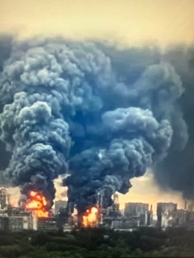 The image depicts a large industrial fire with massive plumes of dark smoke billowing into the sky. The fire is concentrated in the foreground, illuminating the surrounding structures with an intense orange glow. In the background, the silhouettes of buildings and industrial equipment are visible, suggesting an urban or industrial setting. The sky is overcast with a yellowish hue, potentially indicating the time of day or atmospheric conditions. The overall scene conveys a sense of destruction and ongoing emergency. The location is identified as Sof'ino, Russia.