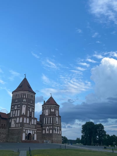 The image shows a view of Mir Castle Complex in the village of Mir, Karelichy District, Grodno Region, Belarus. The castle, a UNESCO World Heritage site, is depicted with its iconic brick towers and distinctive architectural style, set against a backdrop of a blue sky with scattered white and dark clouds. In the foreground, there is a well-maintained lawn and paved pathways. To the right of the castle, a large, lush green tree stands prominently. The overall scene suggests a serene and historical setting.
