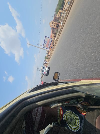 The image depicts a daytime street scene in AlakIa, Nigeria, captured from the interior of a moving vehicle, looking forward and to the right. In the immediate foreground, the upper frame of the vehicle's passenger door and a portion of the side mirror are visible. A person's right shoulder and head, partially obscured, are present in the lower right, wearing a dark garment with a distinctive yellow, red, and white circular pattern.
The main focus is a wide asphalt road, likely a multiple-lane thoroughfare, extending into the distance under a clear blue sky with scattered white clouds. Several vehicles are on the road; a white SUV or car is centrally located further down, with a red vehicle partially visible behind it, and other indistinct vehicles further in the background. Along the right side of the road, a concrete barrier separates the pavement from a pedestrian area. Several small figures are visible on this side, appearing to walk or stand. A tall pole supports a prominent blue and white billboard, which displays partial text including "PROPERTY OF LIFE ASSEMB...". Overhead utility lines are visible against the sky.
In the mid-ground and background, a large, ornate building with a reddish-brown facade, featuring multiple arched windows and distinct architectural details, stands prominently on the right side beyond the road. Hilly terrain or additional structures rise behind this main building in the far distance.
