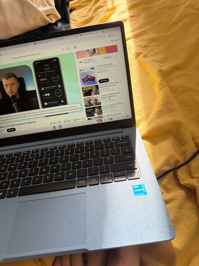 The image shows a light blue laptop on a wrinkled yellow surface, likely a bedspread. The laptop is open, displaying a YouTube page with thumbnails of various videos, including content on investing and finance. The laptop keyboard is visible with the "Intel Celeron" sticker near the top right corner. The screen is lit, and the cursor is visible. A black cord is partially visible in the top right corner of the image, possibly a charging cable. The keyboard is black with gray lettering. Part of a person's leg is visible to the right of the laptop. The image does not depict any location within the city of Pentre, United Kingdom.