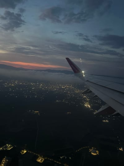The image shows a view from an airplane window during twilight. The airplane's wing is prominently featured in the foreground, extending from the left side of the frame. The wing has the Wizz Air logo printed on its underside in red and white. Below the wing, a city is visible, illuminated by countless lights that create a sparkling tapestry against the dark landscape. In the distance, mountains are silhouetted against the colorful sky, with hues of orange and pink hinting at the setting or rising sun. The sky itself is filled with clouds, some dark and moody, others lighter and tinged with the fading daylight. The overall scene evokes a sense of transition, perhaps the end of a day or the beginning of a night flight, with a feeling of quiet observation and wonder as the plane glides over the sleeping city.