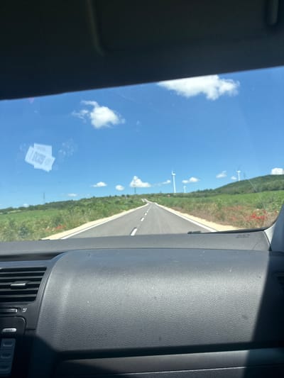 The image is taken from the interior of a vehicle, looking through the windshield. The dashboard is visible in the foreground, with an air vent and part of the infotainment system on the left. On the windshield, there are two white stickers with QR codes.

The view through the windshield shows a paved road stretching into the distance. The road is bordered by green fields, with scattered red flowers, likely poppies. In the background, rolling hills are covered in green vegetation and dotted with several wind turbines. The sky is bright blue with a few scattered white clouds. The scene suggests a sunny day in a rural area. Based on the context provided about Suvorovo, Bulgaria, this could be a road leading towards or away from the city, with the surrounding landscape characteristic of the region.