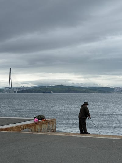 An overcast, grey sky hangs over a calm, grey sea in this full vertical shot. The foreground features an asphalt surface leading to a concrete pier with rusted metal edges. On the pier, a pile of dark clothing and a bright pink object are visible. In the mid-ground, a lone figure, a man wearing a hat and dark clothing, stands facing the water, holding a fishing rod. His posture suggests he is fishing. In the background, across the water, a large cable-stayed bridge with a prominent central pylon dominates the horizon. Beyond the bridge, a green, hilly landscape is dotted with buildings, indicating a city. A cargo ship is visible in the water between the shore and the bridge, and further in the distance, more city structures can be seen along the coastline. The overall impression is one of a quiet, somewhat somber day by the sea in a coastal urban environment, possibly Russky Island, Russia, given the distinctive bridge architecture.