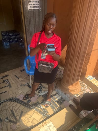 The scene depicts a transaction taking place at a Moniepoint agent location, identified by a sign displaying "MONIEPOINT" and "9067449912 OKORO CHIKA MARYJANE," situated in Aguata, Nigeria.

A young adult female stands centrally, facing downwards, focused on her hands. She has dark skin and braided hair. Her attire consists of a red short-sleeved v-neck t-shirt, a black knee-length skirt, and black slide sandals with "CELINE" branding. A brown patterned shoulder bag is worn across her body. In her left hand, she holds a light blue Moniepoint Point-of-Sale (POS) device. Her right hand manipulates a stack of Nigerian Naira banknotes, seemingly inserting them into or withdrawing them from the device. Her mouth is slightly open.

To the lower right, the lower legs and feet of a second individual, barefoot, are partially visible.

The immediate environment features a tiled floor composed of irregularly shaped, multi-colored tiles in shades of beige, black, and white. Strong natural light creates distinct shadows on the floor and a large, fluted, light brown architectural column on the right side of the frame. To the left, an open doorway reveals a dimly lit interior with numerous stacks of bottled water, and a blue plastic chair is partially visible near the doorway. The background wall is an unadorned light brown/orange color.