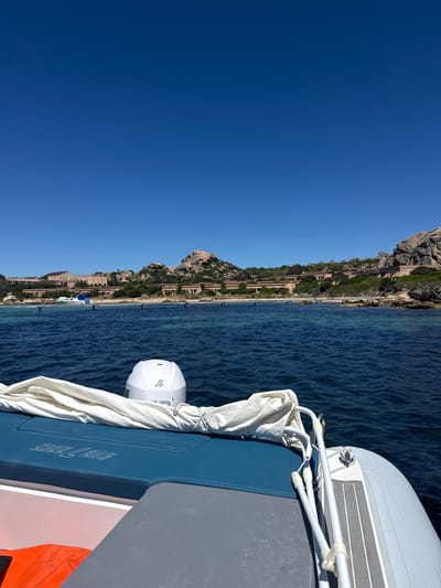 This is a photograph taken from the perspective of someone on a boat, looking towards the shore under a clear blue sky. The foreground shows the interior of a white inflatable boat with blue and grey seating, and an orange life vest is visible on the left. A white outboard motor is mounted at the back of the boat. The water is a deep blue, with some choppiness. In the middle ground, several wooden posts are visible in the water, suggesting a mooring area or a shallow area. Beyond the water, a shoreline is lined with buildings, appearing to be a resort or residential complex, constructed in a style that blends with the natural landscape. The buildings are tan and spread out along the coastline, with a backdrop of rocky, tree-covered hills. The overall scene depicts a sunny day in a coastal location. Given the architectural style and the presence of the sea, this scene is consistent with locations in or around the La Maddalena archipelago in Italy, known for its beautiful coastline and islands.