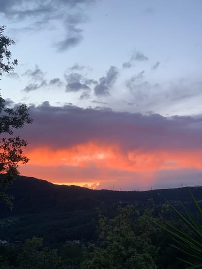 The image shows a twilight or sunset view in Can Güell, Spain. A sharp, dark, tree-covered mountain ridge line dominates the right side of the frame, with a glimpse of buildings and power lines along the crest. To the left of the ridge, vibrant orange and pink hues illuminate thick, layered clouds, suggesting the sun is setting behind the mountain. On the left side of the image, the sky shifts to a pale blue-gray with scattered clouds. In the foreground, branches of trees obscure the bottom of the view.