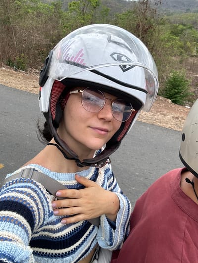 The image shows a woman wearing a white helmet with a clear visor and round glasses, sitting on what appears to be a motorcycle. She is wearing a blue and white striped open knit sweater, and a seatbelt is visible across her torso. Another person wearing a helmet and a red shirt is partially visible to the right of the woman. The background reveals a road, vegetation-covered terrain, and distant hills. The location is Hàm Thuận Bắc, Vietnam.