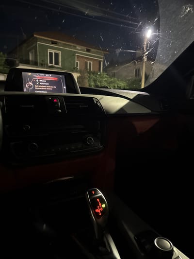 The image shows the interior of a car at night. The dashboard display shows the car's infotainment system, with options for "iPhone" and "AUX front." The gear shifter is visible, illuminated with the letter "P" for park. Outside the car, a house with green walls and a brown roof is visible, along with a street lamp and power lines. The scene is set in Zverino, Bulgaria, as indicated by the metadata. There are no people or specific actions depicted in the image. The environment is dark and somewhat cluttered with dust or debris on the windshield.