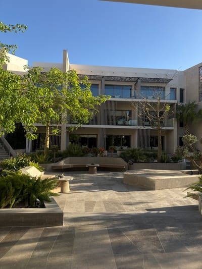 The image shows a building with a modern architectural design, featuring multiple stories with balconies and decorative metalwork. There are palm trees and other greenery interspersed within the building's structure, adding a natural element to the urban environment. In the foreground, a tiled pathway leads towards the building, with benches and landscaping elements along the sides. The overall setting is sunny with a clear blue sky, suggesting a warm climate. The location is El Gouna, Egypt, based on the architecture and landscaping style. The scene appears to be a courtyard or open space within a resort or residential complex.