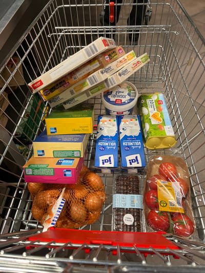 A shopping cart contains assorted groceries. There are packages of Wagner's frozen pizzas, including salami and mozzarella flavors. Also present are tubs of Joghurt mild, cartons of ja! Vollmilch (whole milk), boxes of Kölln muesli (including Knusper Honig-Nuss), and a bag of onions. Additional items include a package of REWE "10 frische Eier" (10 fresh eggs), a pack of tomatoes, and a package of chocolate items with an expiration date of 30.08.2026.
