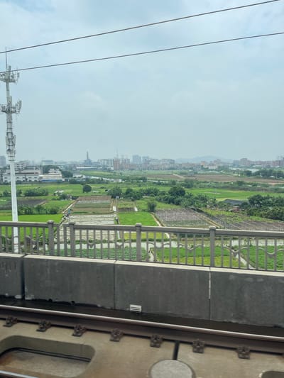 The image captures a view from a moving train or elevated platform, looking out over a sprawling landscape. In the foreground, a concrete structure with a metal railing separates the viewer from the scene beyond. The railing features an intricate, repeating pattern of white arches against a backdrop of lush green fields and water channels. Beyond the railing, a diverse mix of residential buildings and agricultural land stretches into the distance. Tall apartment buildings, some under construction with visible cranes, are interspersed with smaller houses and what appear to be rice paddies or other cultivated fields. Numerous trees dot the landscape, adding to the green expanse. In the far distance, a hazy mountain range is visible under a bright, overcast sky. The overall impression is one of a developing urban fringe, where the city meets the countryside. The time of day appears to be daytime, with the sky indicating it could be a cloudy or hazy day. The presence of power lines and communication towers in the lower left corner suggests an urban or semi-urban environment. The photo's orientation is slightly tilted, giving a dynamic feel to the perspective.