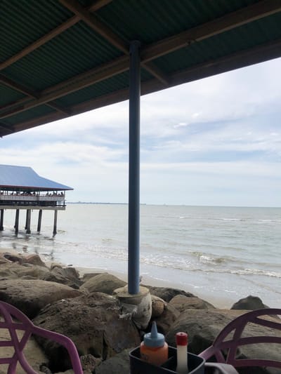 This media file depicts a beachside scene in Lancok, Indonesia. The viewpoint is from under a corrugated metal roof structure, with wooden beams supporting it. In the foreground, a large, grey, cylindrical pole runs horizontally across the frame, obscuring a portion of the view. To the right of the pole, a rocky embankment lines the beach.
The ocean is visible, with gentle waves lapping at the shore. In the distance, a pier extends into the water, with a building on stilts. The sky is overcast, with scattered white clouds, suggesting it may be daytime, possibly late morning or early afternoon.
On the right side of the image, a purple chair is partially visible, and a dark container holds a red-capped bottle and an orange-capped squeeze bottle, possibly condiments. The overall atmosphere appears relaxed and casual, suggesting a beachside dining or recreational area. There is no visible text in the image.