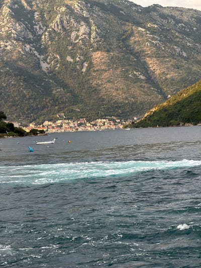 This is a medium shot photograph taken from a body of water looking towards a coastal town situated at the base of a steep, mountainous incline. The water in the foreground is choppy with significant white foam and a distinct band of turquoise-colored turbulent water running horizontally across the frame.
In the middle ground, the dark blue water is calmer, with a white motorboat visible in the center. Two buoys, one blue and one yellow, are also present in the water. To the left of the boat, a white structure, possibly a building, is partially visible on the shore.
The background features a densely populated coastal town with buildings exhibiting red-tiled roofs and light-colored facades. The town is nestled against a large, rugged mountain that dominates the upper portion of the image. The mountain's slopes are covered with sparse vegetation and rocky outcrops. The lighting suggests it is either early morning or late afternoon, as the sun casts shadows across the mountain.
The scene appears to be in or near Đurići, Montenegro, given the characteristic landscape of the Bay of Kotor, which features steep mountains meeting the sea and picturesque coastal towns. There are no people clearly visible in the image.