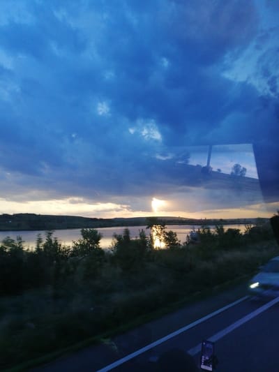 This is a photograph of a sunset over a body of water, likely a river or lake, with a landscape of hills and trees in the background. The sky is filled with dark blue and gray clouds, with a bright patch of sunlight breaking through near the horizon. The reflection of the sky and landscape is visible on the water's surface. In the foreground, there is a road with a car driving on it, its headlights illuminated. There is also a smartphone mounted on a stand on the road. The image appears to be taken from inside a vehicle, possibly a bus or car, given the reflection in the window. The location is identified as Pocoleni, Romania.