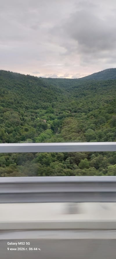 The image depicts a panoramic view of a densely forested mountainous landscape, likely taken from a moving vehicle. The foreground features two metallic guardrails, indicative of a road or highway. Beyond the guardrails, a vast expanse of evergreen trees covers the rolling hills, with a narrow dirt path or stream visible winding through the foliage. The sky is overcast with heavy, grey clouds, suggesting a cloudy or potentially rainy day. In the bottom left corner of the image, text indicates the device used to capture the photo, "Galaxy M52 5G," and the date and time, "9 юни 2026 г. 06:44 ч." (June 9, 2026, 06:44). The overall impression is one of a natural, remote setting.