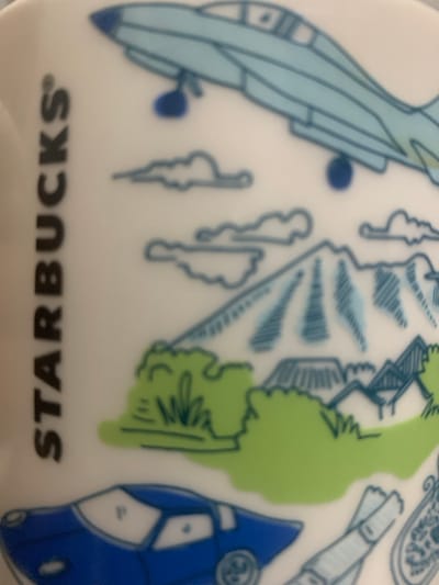 The image shows a close-up of a white mug with a cartoon-style illustration. On the left side, the word "STARBUCKS" is printed vertically in black. The illustration depicts various elements: a light blue jet in the upper portion, stylized clouds, a mountain with blue hatching, green grassy hills, and some buildings that appear to be houses with blue roofs. In the lower part of the image, a blue and white sports car is visible. To the right of the car, there is a drawing of a pair of ski poles and a paisley-like design. There is no indication of the city of Angeles, Philippines in the image.