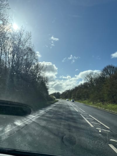 The image displays a road scene in West Felton, United Kingdom, captured from the interior of a vehicle looking forward through a windshield. The scene is illuminated by bright sunlight under a partially cloudy blue sky, with the sun visible in the upper left and generating glare on the windshield. A two-lane asphalt road, dark grey in color, extends into the distance, curving slightly to the right. White dashed lane lines are visible, along with a distinct segment of white rectangular markings on the right side of the road. Dense deciduous trees, appearing mostly bare or with nascent foliage consistent with late winter or early spring, line both sides of the road, forming a natural corridor. Three vehicles are visible ahead on the road, traveling in the same direction as the vantage point; they appear as indistinct shapes with illuminated headlights, with one vehicle appearing dark and another grey/silver. The immediate foreground shows the textured pattern of the vehicle's windshield and a dark, indistinct component of the dashboard. No individuals are discernible.