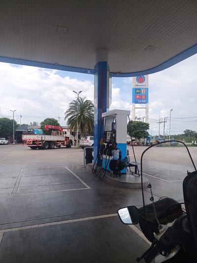 The image depicts a gas station in Tha Ruea, Thailand. A man in a blue and white shirt is sitting next to a gas pump. In the background, there's a large truck with a red crane mounted on it, and another vehicle is parked nearby. A PTT Station sign is visible in the background, along with a 7-Eleven logo. The scene is partially obscured by a motorcycle windshield in the foreground. The gas station is located on a paved area with road markings. Overhead, a gas station canopy provides shelter.