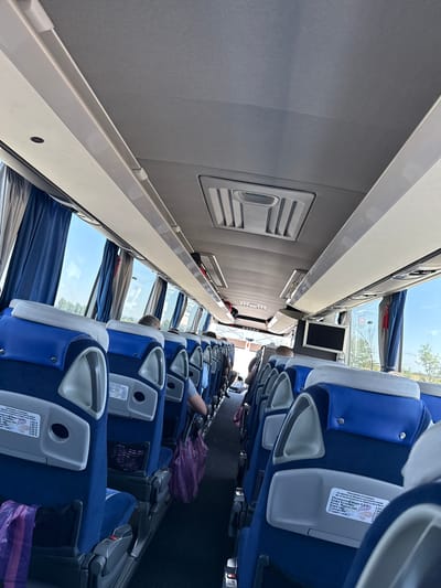 The image shows the interior of a bus, looking forward from a seat in the back. The seats are blue with gray headrests and have a typical bus seat design with individual armrests. Some seats are occupied by passengers, who are mostly seen from the back or side. There are blue curtains drawn over the windows, revealing a bright sky and some trees outside. The bus has overhead compartments and lighting. A small television screen is mounted on the ceiling in the middle of the bus. There are price lists visible on the seat backs, indicating items for sale on the bus, such as mineral water, coffee, beer, Fanta, and tea. The environment appears to be a sunny day, and the bus is likely in motion, as suggested by the slight blur of the exterior. The location is identified as Belozem, Bulgaria.