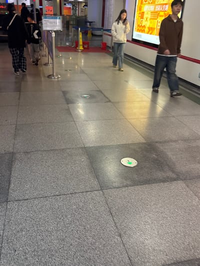 An indoor space with tiled flooring features several individuals. Two people stand near a wall with an advertisement. A woman in a light-colored jacket and jeans is slightly behind a man in a brown shirt and jeans. Further down, another group is present. Circular markers with green arrows are embedded in the floor, suggesting a directional system. Metal crowd-control barriers are also visible.