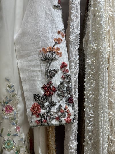 This is a close-up, medium shot of three garments hanging next to each other. The garment on the left is white with intricate floral embroidery in various colors, including pink, blue, and green, adorned with sequins and beads. The central garment is also white and features a dense, textured lace with attached pearls and sequins. The garment on the right appears to be a cream-colored lace with a similar pearl and sequin embellishment, but with a more open pattern. The lighting is soft and diffused, highlighting the delicate details of the embellishments. The background is dark and indistinct, suggesting the garments are displayed indoors, likely in a retail or studio setting. There are no people or animals visible in the image, and no indication of a specific location within Tanjungmeru, Indonesia.