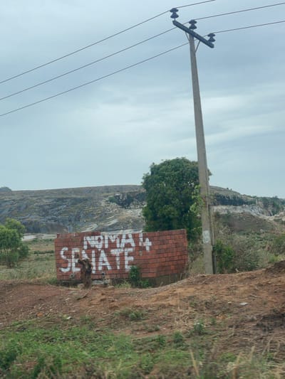 A brick wall stands beside a dirt road at the base of a rocky slope, with graffiti "Skate Novam" painted on the wall. A utility pole is positioned on the left side of the image. The scene is located near the quarry in Kujama, Nigeria.