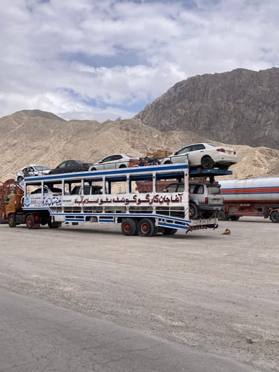 A large, custom-built, multi-level car carrier truck is prominently featured, parked on a dirt and gravel roadside. The truck, predominantly white with blue accents and an elaborate metal framework, is heavily laden with vehicles. Visible on its different tiers are at least three cars: a white sedan on the top level, a gray SUV below it, and another white car towards the front. Behind this carrier, part of a second large truck, possibly a tanker or another cargo vehicle adorned with traditional South Asian truck art in red, white, and blue, is partially visible.
The scene is set against a backdrop of imposing, rugged mountains with a dry, rocky appearance, indicating an arid or semi-arid mountainous region. The sky is partly cloudy, suggesting it is daytime. On the ground beside the car carrier, a small, light brown animal, possibly a dog or fox, is visible.
The car carrier features discernible text on its side, primarily in Urdu, which reads:
- "Ν.Τ.Ν. 8.2975591" (likely a registration or identification number)
- "آغا جان کارگو گوشه و اسلام آباد" (translates to "Agha Jaan Cargo, Gosha and Islamabad," indicating a cargo company and possibly destinations)
- "قاہر گل بھائی ہوں اسلام کو 24" (possibly a name "Qahir Gul Bhai" and further route or identification details related to "Islamabad," e.g., "Qahir Gul Bhai, from Islamabad, Route 24").
The image captures a scene of overland transport in a mountainous terrain, with the custom modifications of the truck and the presence of Urdu text strongly suggesting a location in Pakistan.