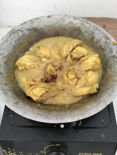 Chicken pieces simmer in a pale yellow sauce within a large, weathered wok. The wok sits on a black Euro Gas stove, its surface marked with wear. The sauce bubbles gently around the chicken, indicating active cooking. The scene conveys a simple, home-cooked meal in progress.