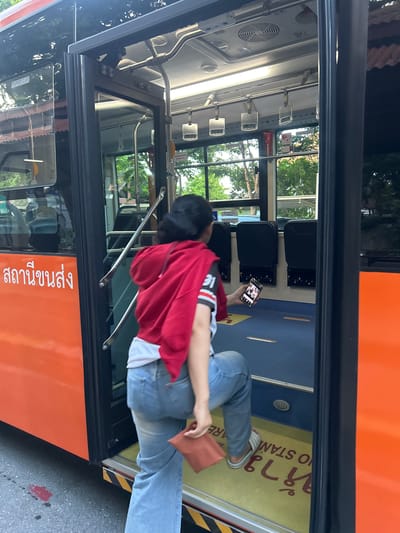 A young Thai woman is captured from behind, stepping onto a bus in Tha Khlong Town Municipality, Thailand. She is wearing light blue denim jeans, a white t-shirt with black and red stripes on the sleeves, and a red hooded sweatshirt draped over her shoulders. In her right hand, she holds a smartphone, and in her left, a small brown pouch or wallet. The bus is orange with Thai writing on its side. The bus interior features blue flooring with yellow and red warning stripes near the entrance, and rows of black seats. Overhead, metal handrails are visible. The scene appears to be outdoors, with trees and buildings visible through the bus windows.
