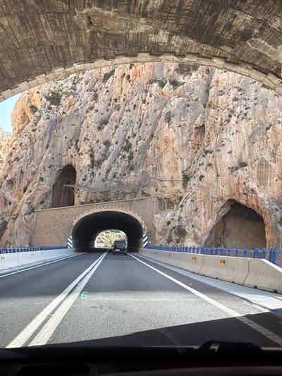 The image shows a view from inside a vehicle looking out at a road leading into a tunnel. The tunnel entrance is a large archway constructed from stone, built into the side of a rocky mountain. The mountain face is rugged and appears to be made of light-colored rock, with some vegetation growing in crevices. There are two large cave-like openings in the mountain face above and to the sides of the tunnel. The road itself is paved asphalt with double white lines. A dark-colored van is visible driving into the tunnel ahead. The vehicle's dashboard is visible in the foreground, and a green digital display, possibly a speed indicator, shows the number "6". The road is bordered by concrete barriers with blue railings and orange reflectors. The scene suggests a drive through a mountainous coastal area, consistent with the coastal region of Altea, Spain.