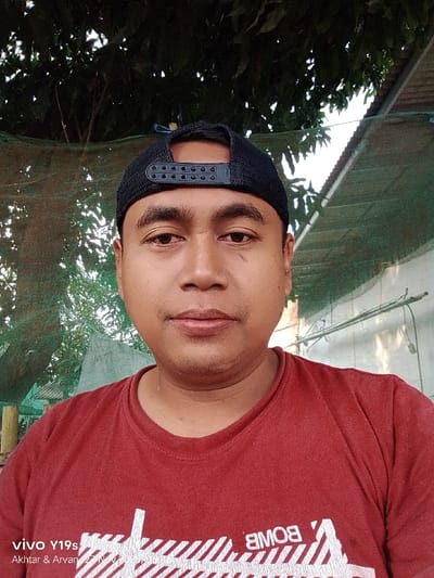 A man in a red t-shirt and black baseball cap is captured from the chest up. His skin is tan, and he looks directly at the camera. The background is slightly blurred and appears to be outdoors, with trees and a building partially visible. The bottom left corner contains text indicating the device model and names.