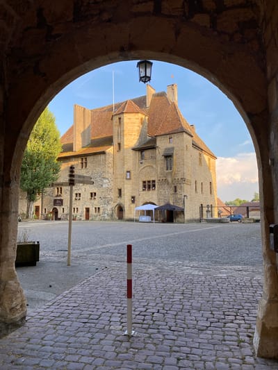 An arched stone entryway frames a view of a historic stone building with a red-tiled roof under a clear blue sky. A flag flies from the building's highest point. In the foreground, a cobblestone plaza stretches out, with a signpost indicating directions. Two temporary tents, one white and one black, are set up in front of the building. A red and white striped bollard stands on the cobblestones. To the right, a dark blue car is parked behind a metal fence. The environment appears to be a public square in a historic European town. There are no people clearly visible in the scene.