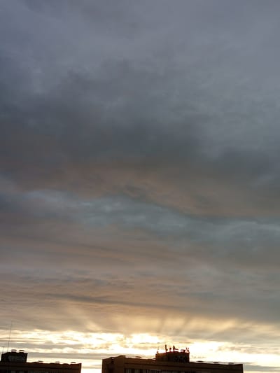 This media file depicts a cloudy sky over Usol'e, Russia, likely at dusk or dawn due to the warm, glowing hues in the lower portion of the sky. The clouds are varied in texture and color, ranging from dark, ominous grays at the top to lighter, wispy formations tinged with orange and yellow closer to the horizon. Streaks of light break through the clouds, creating a dramatic effect.
In the foreground, the tops of two buildings are visible. The building on the left has graffiti on its facade and a prominent antenna or lightning rod. The building on the right is also topped with structures, possibly related to telecommunications or HVAC systems. The architecture suggests an urban or industrial setting.
The overall mood is somewhat somber, characteristic of overcast weather, but the colorful light at the horizon adds a touch of beauty and hope. There is no visible activity or people in the frame, focusing the viewer's attention entirely on the interplay of light and cloud formations. The text "TGT" and other illegible graffiti are visible on the left building.