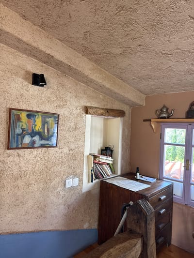 This image depicts an interior scene within a building, characterized by textured walls, likely stucco or a similar material. The color palette is predominantly earthy tones of beige and brown. The lighting appears to be a mix of natural light filtering in from a window and artificial light.
The main architectural features include a staircase leading upwards, a window with a lilac frame through which a blurred outdoor scene can be observed, and a built-in niche or shelf. In the niche is a collection of books and other objects.
Objects of interest are the silver teapot mounted near the window, a framed painting hung on the wall, an electrical spotlight fixture, and a pair of electrical switches. There is a piece of furniture, perhaps a chest of drawers, and a rough-hewn wooden beam in the corner.
The environment does not show any indication of where the image might be located, or any people present.