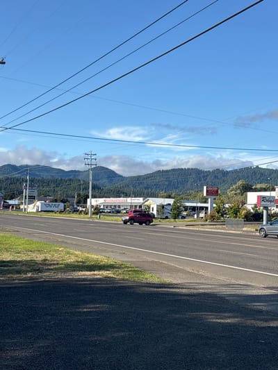 A roadside scene captures a highway adjacent to buildings and signs, potentially commercial establishments. A red vehicle is parked near a building marked with "Camping." Mountains are partially visible in the backdrop, flanked by a sky with power lines. The day appears clear, with a few clouds scattered across the blue sky. The composition emphasizes the roadside infrastructure and landscape.