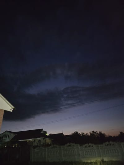 The image depicts a twilight scene in Moscovei, Moldova, featuring a residential area with houses and a decorative concrete fence. The sky is dark and cloudy, with hints of purple and orange near the horizon, suggesting the end of sunset. In the foreground, a part of a house with a white roof and brown brick wall is visible on the left. Further back, several houses are silhouetted against the sky. A decorative concrete fence with arched patterns runs across the middle ground. Trees are visible behind the fence. Power lines are strung across the sky. The lighting is dim, indicating it is likely evening or pre-dawn. No people or animals are present in the frame.
