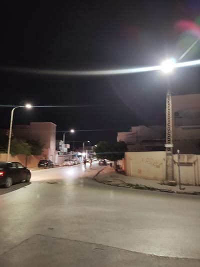 The image depicts a street scene at night in Raoued, Tunisia. A black car is driving on the left side of the road, with its headlights on. Several other vehicles are parked along the right side of the street, and some streetlights illuminate the scene. Buildings line the street, with one notable structure on the right featuring a tall utility pole with a ladder-like design. The road surface appears to be paved, and there are areas of shadow and light due to the artificial lighting. A person or people can be seen in the distance on the right side of the street. The sky is dark, indicating nighttime.
