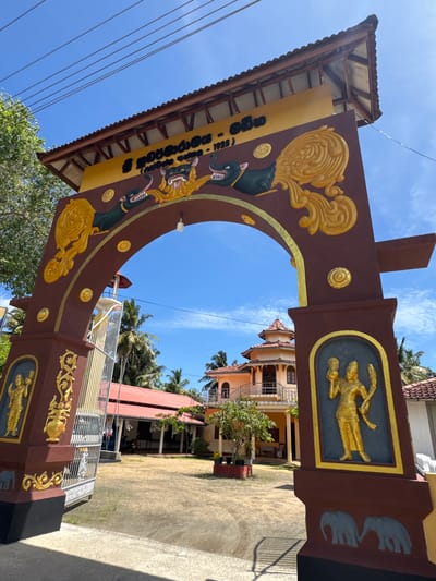 An ornate, traditional entrance archway, viewed from a slightly upward angle, stands prominently under a bright blue sky with scattered clouds. The archway is primarily dark brown, richly decorated with gold-colored relief sculptures and patterns. Along the curve of the arch, two dark green mythical creatures, possibly Makara, with visible fangs and tongues, are depicted exhaling elaborate golden scrollwork. Several circular gold medallions are also interspersed on the arch.

The two main pillars supporting the arch feature rectangular recessed panels containing golden relief sculptures of two human-like figures each, possibly deities or traditional dancers. Further down the right pillar, smaller grey elephant motifs are visible. A yellow banner hangs on the left side of the archway, below a dark-tiled roof. The text on the banner is in Sinhala script and reads "ශ්‍රී ගොඩගම වල්ගම" (Sri Godagama Walgama), "පුරාණ විහාරය" (Ancient Temple), and the year "(1925)", suggesting it is the "Sri Godagama Walgama Ancient Temple, Established 1925."

Beyond the archway, traditional Sri Lankan buildings with red-tiled roofs and light-colored walls are visible, surrounded by green foliage and palm trees. The ground appears to be a paved courtyard leading into the complex. The bright sunlight indicates it is daytime, likely late morning or early afternoon, with clear weather. Power lines are visible against the blue sky on the left side. The scene depicts the entrance to a culturally and historically significant site in Walgama, Sri Lanka.