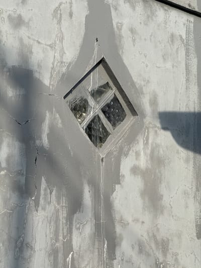 A diamond-shaped window with textured glass panes is set into a rough, gray stucco wall. The wall exhibits signs of age and weathering, with visible cracks, water stains, and patches of lighter and darker gray paint, suggesting a recent repair or application of a sealant. The window is framed by a slightly raised border, also finished in gray. Sunlight casts a shadow of an unseen object onto the right side of the wall. The scene appears to be an exterior wall of a building, likely residential or commercial, in Sukawati, Indonesia. There are no people or active events depicted in the image.