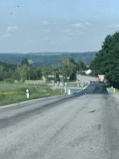 This is a blurry, overexposed, daytime photograph taken from the perspective of a vehicle moving along a rural road in Šternov, Czechia. The road curves to the right and leads towards a junction with traffic signs. In the distance, a small red truck is visible. The surrounding landscape is characterized by rolling hills covered in green trees and fields. The sky is a pale, washed-out blue with hints of clouds. The overall impression is one of a bright, overcast day. The image quality is poor, with numerous small dark spots scattered across the frame, likely dust or imperfections on the lens or sensor. The scene is peaceful and depicts a typical rural setting in the Czech countryside. There is no visible text in the image.