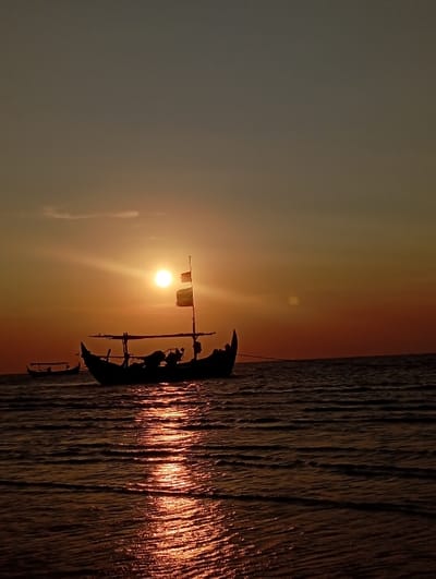 The image displays a sunset scene over a body of water in Tritunggal, Indonesia. A traditional Indonesian fishing boat, silhouetted against the setting sun, is centrally positioned in the mid-ground. The boat features a high, curved bow and stern, a central framework structure, and a mast with a flag or banner attached. An engine or equipment is faintly discernible within the boat's hull. The sun, a bright orange-yellow orb, is low on the horizon, casting an intense, shimmering reflection path that extends across the rippled water surface towards the foreground. The sky exhibits a color gradient, transitioning from bright orange and yellow at the horizon to a darker grey-blue overhead. A second, smaller boat is visible in silhouette in the distant background to the left. No individuals are visible.