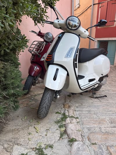 A white and burgundy vintage Vespa scooter sits parked beside a stone wall in the city of Grad Rovinj, Croatia. The white scooter is positioned on top of the burgundy scooter. Both scooters face away from the wall. The scene includes views of the scooters' tires, headlights, mirrors, and seats. The scooters are parked near some greenery. The building is in the background.