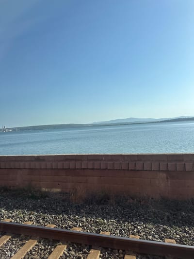 The image depicts a tranquil waterscape with a clear blue sky. In the foreground, a section of a brick wall runs horizontally across the frame, partially obscuring a railway track with gravel ballast. Beyond the wall, a wide expanse of calm blue water stretches towards the horizon. In the distance, a low-lying landmass with rolling hills is visible, dotted with industrial structures and vegetation. The overall impression is one of a serene, natural setting juxtaposed with man-made elements. The specific location in Bages, France, is not identifiable from the visual information provided in the image.
