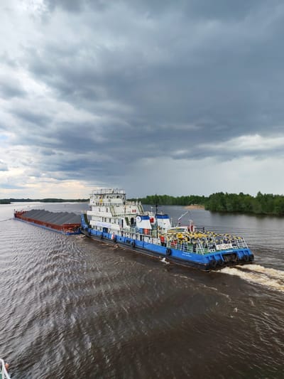 A river scene in what appears to be a cloudy environment. A blue and white tugboat, "OT-2410," is pushing a barge laden with dark material, possibly coal or gravel. The scene is likely near a riverbank, as indicated by the tree line visible on the right side of the frame. No people are clearly visible. Given the location provided, this is presumably happening in or near Kargulino, Russia. The water is dark and disturbed by the movement of the vessels.